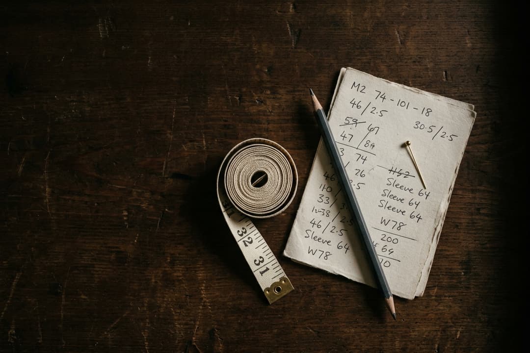 A cream tape measure coiled beside a folded handwritten measurement card and pencil on a dark wooden tailor's bench.