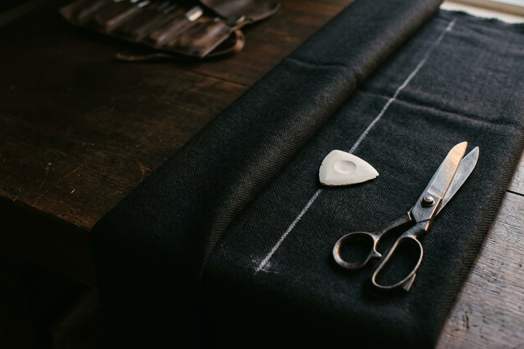 A white chalk line drawn across dark herringbone suiting cloth with steel tailor's shears resting on the fabric.